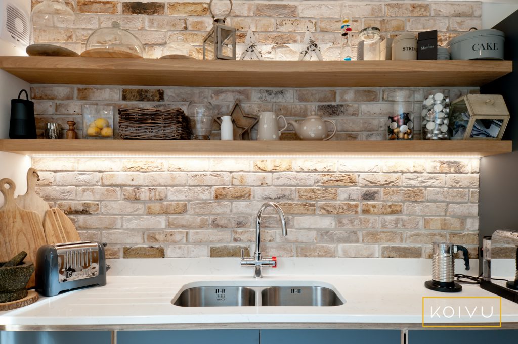 Brushed stainless steel sink inlaid in white worktop. Backed by stone feature wall with open oak shelves.  By Koivu.