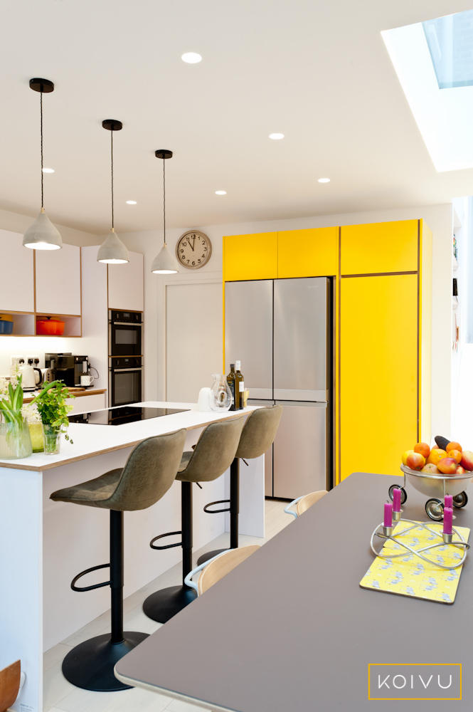 Kitchen island with breakfast bar in a white and yellow kitchen.  Tall, bespoke  yellow units built to house American style fridge / freezer. By Koivu.