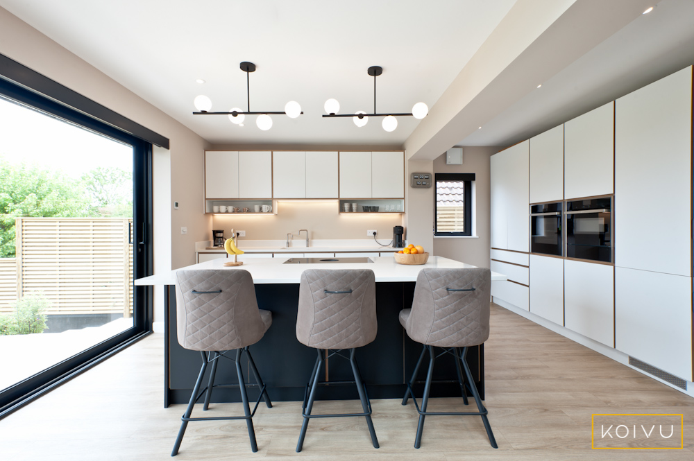 View of white and black kitchen in Whitstable. with island including breakfast bar for 3 seats. To the right hand side there is a long run of tall units including oven housing. By Koivu.