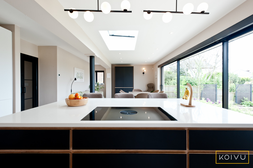 View of this kitchen in Whitstable, fitted in a new large extension. There is a black plywood island in the foreground with a white worktop and hob. In the background you can see the bespoke drinks cabinet made by Koivu. 